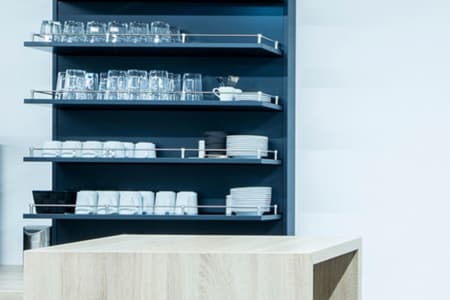 Minimalistische Küchenregale mit ordentlich angeordneten Gläsern, Bechern, Schüsseln und Tellern über einer hellen Holztheke. Der Hintergrund ist klar und modern und betont Ordnung und Schlichtheit.