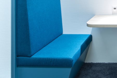 Eine Nahaufnahme einer blau gepolsterten Sitzbank neben einem hellen rechteckigen Tisch, vor einer weißen strukturierten Wand mit dunklem Teppichboden.
