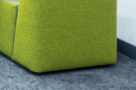 Nahaufnahme der unteren Ecke eines modernen grün gepolsterten Sessels oder Sofas auf einem grauen Teppichboden neben einer weißen strukturierten Wand mit einer dunklen Fußleiste.