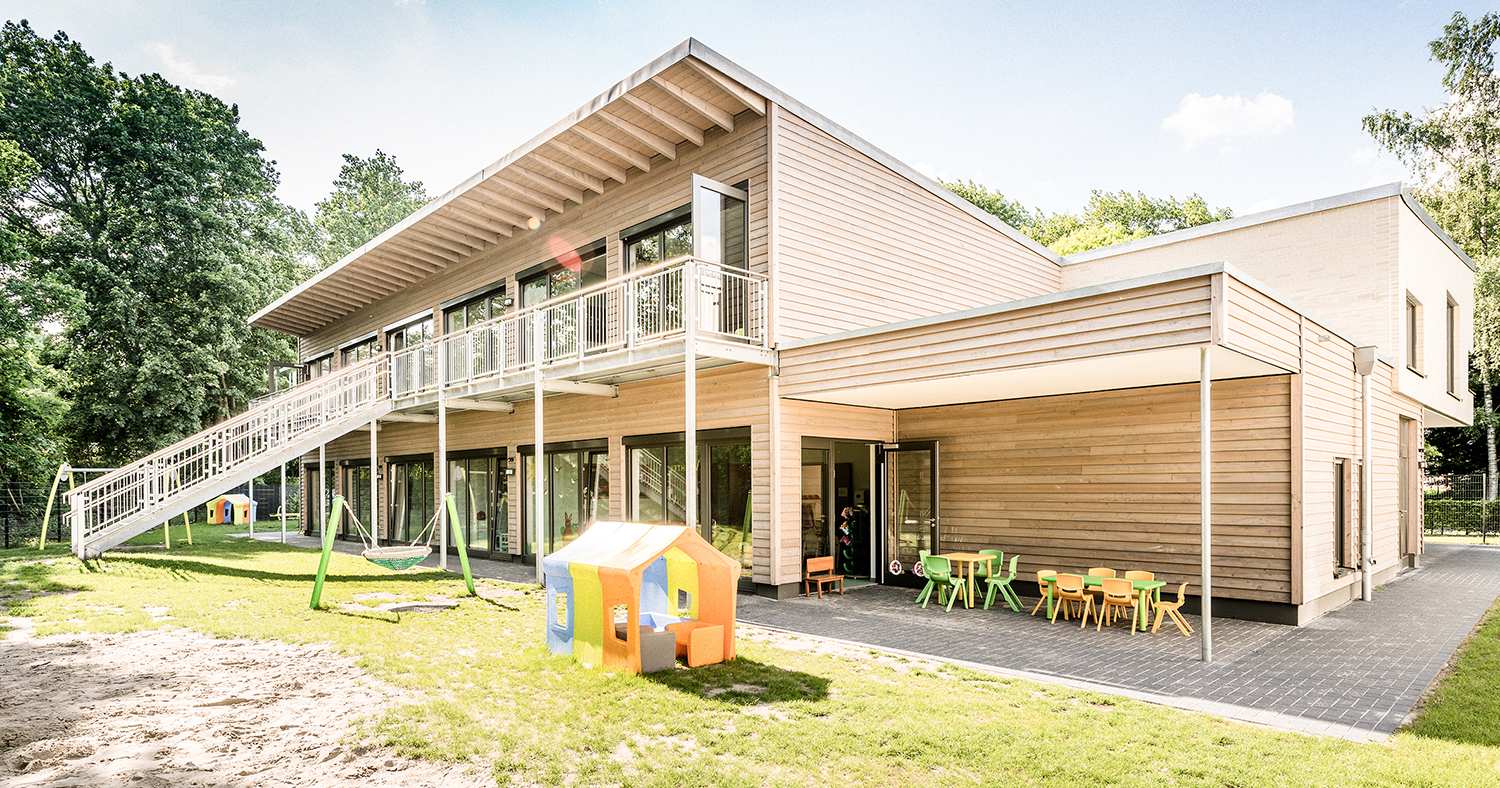 Modernes zweistöckiges Gebäude mit Holzvertäfelung, großen Fenstern und einer Außentreppe. Es gibt einen Spielplatz mit Schaukel, Spielhaus und bunten Tischen und Stühlen auf einer Terrasse, umgeben von Bäumen und Gras.