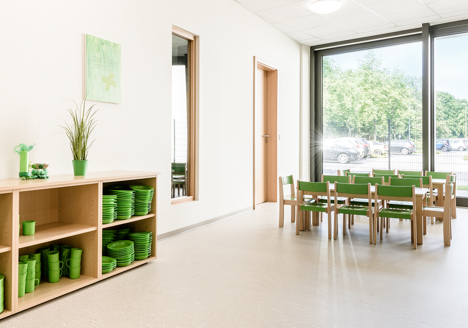Helles Klassenzimmer mit grünen Tellern und Tassen auf Regalen, grünen Stühlen um kleine Tische, großen Fenstern mit Blick auf Bäume und geparkte Autos draußen und einer Pflanze und einem Kunstwerk an der Wand.