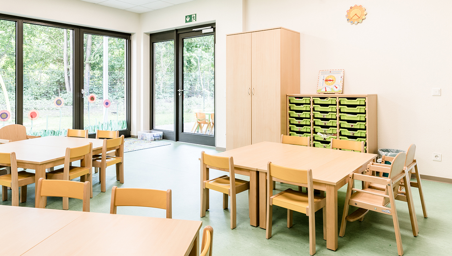 Ein heller, aufgeräumter Klassenraum mit hellen Holztischen und -stühlen, Schränken mit grünen Behältern, großen Fenstern und fröhlicher Dekoration. Der Raum ist von Sonnenlicht durchflutet und bietet Blick auf den grünen Außenbereich.