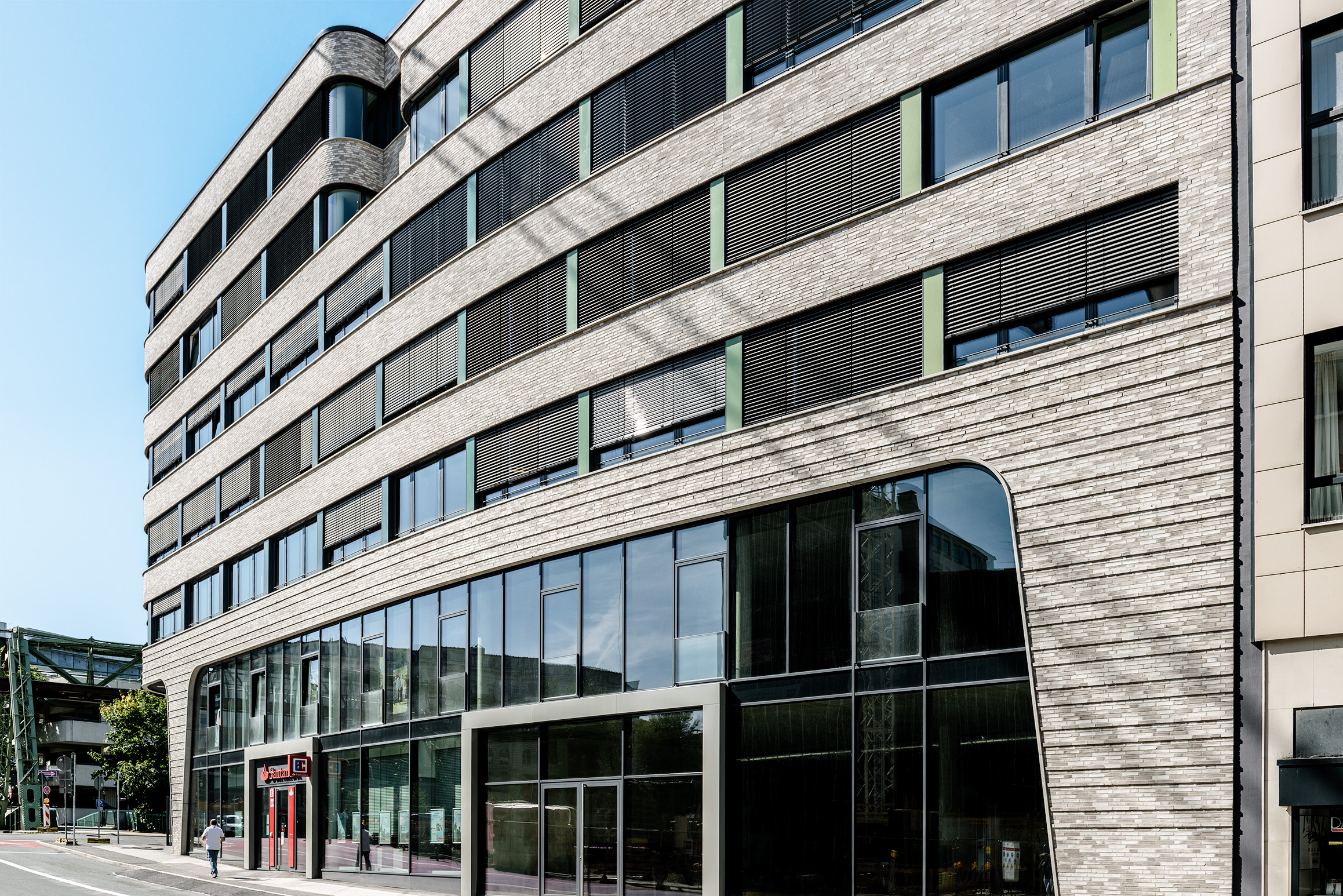 Ein modernes mehrstöckiges Bürogebäude mit großen Fenstern, horizontalen Linien und heller Backsteinfassade, fotografiert aus einem niedrigen Winkel an einem sonnigen Tag. Im Erdgeschoss ist ein rotes Eingangsschild zu sehen.