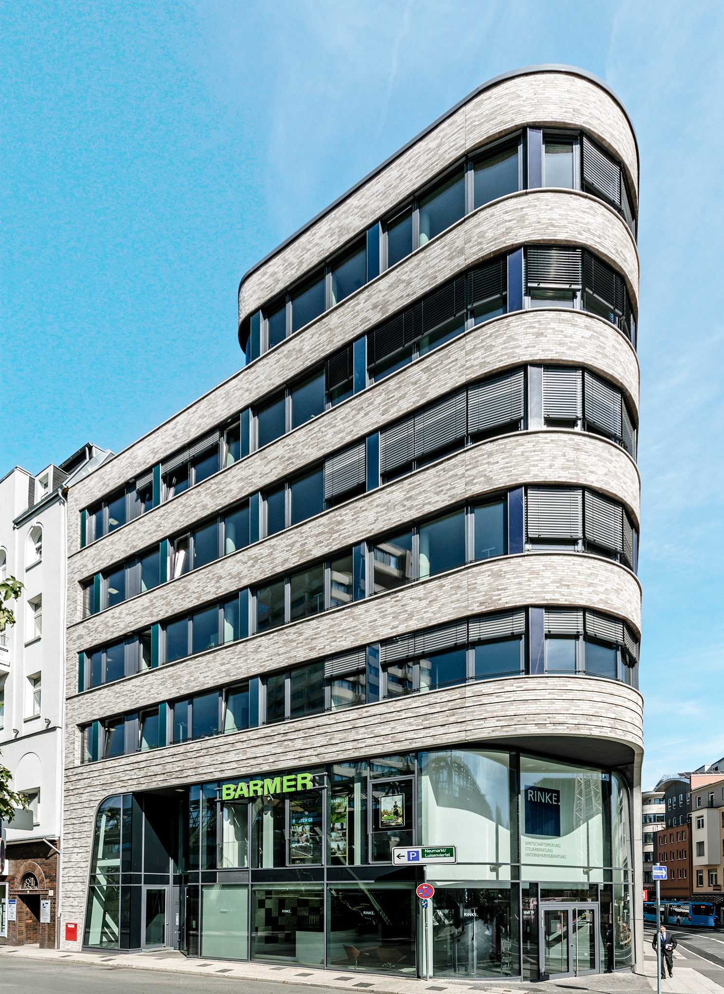 Modernes mehrstöckiges Bürogebäude mit abgerundeten Ecken, großen Glasfenstern und heller Backsteinfassade. Im Erdgeschoss sind die Schilder der Firmen BARMER und RINKE zu sehen. Der Himmel ist klar und blau.