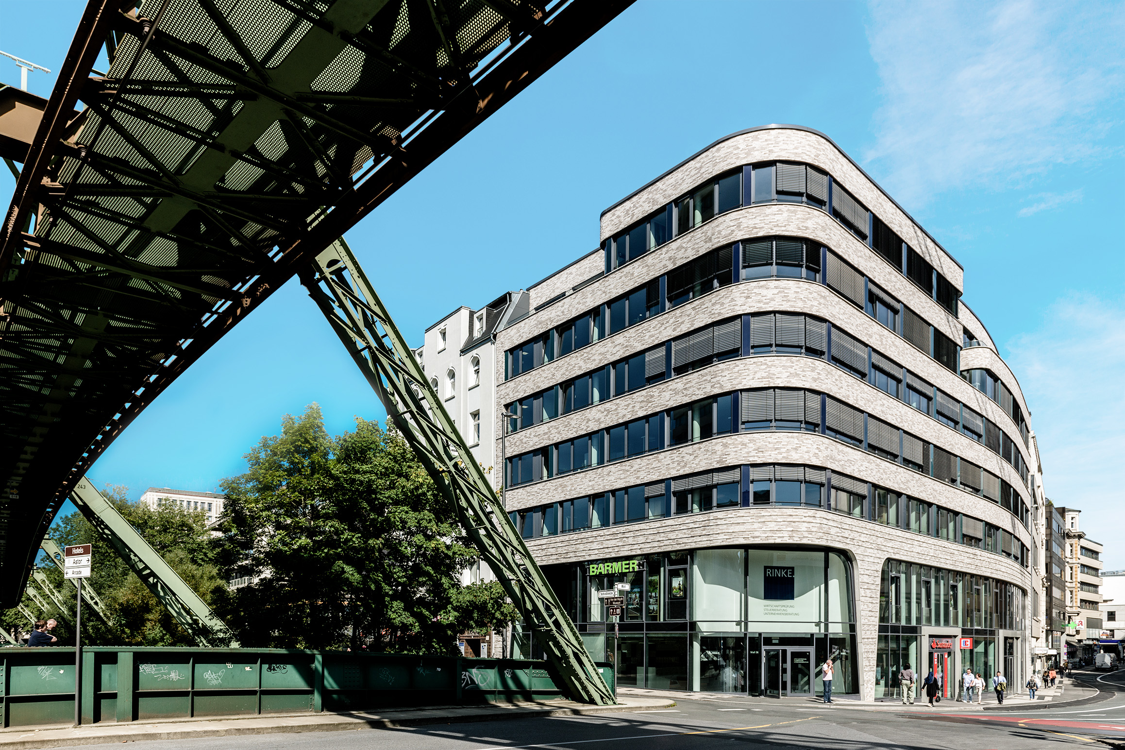 Ein modernes, mehrstöckiges Bürogebäude mit großen Fenstern und abgerundeten Ecken steht neben einer Einschienenbahnstrecke unter einem klaren blauen Himmel auf einer Stadtstraße.