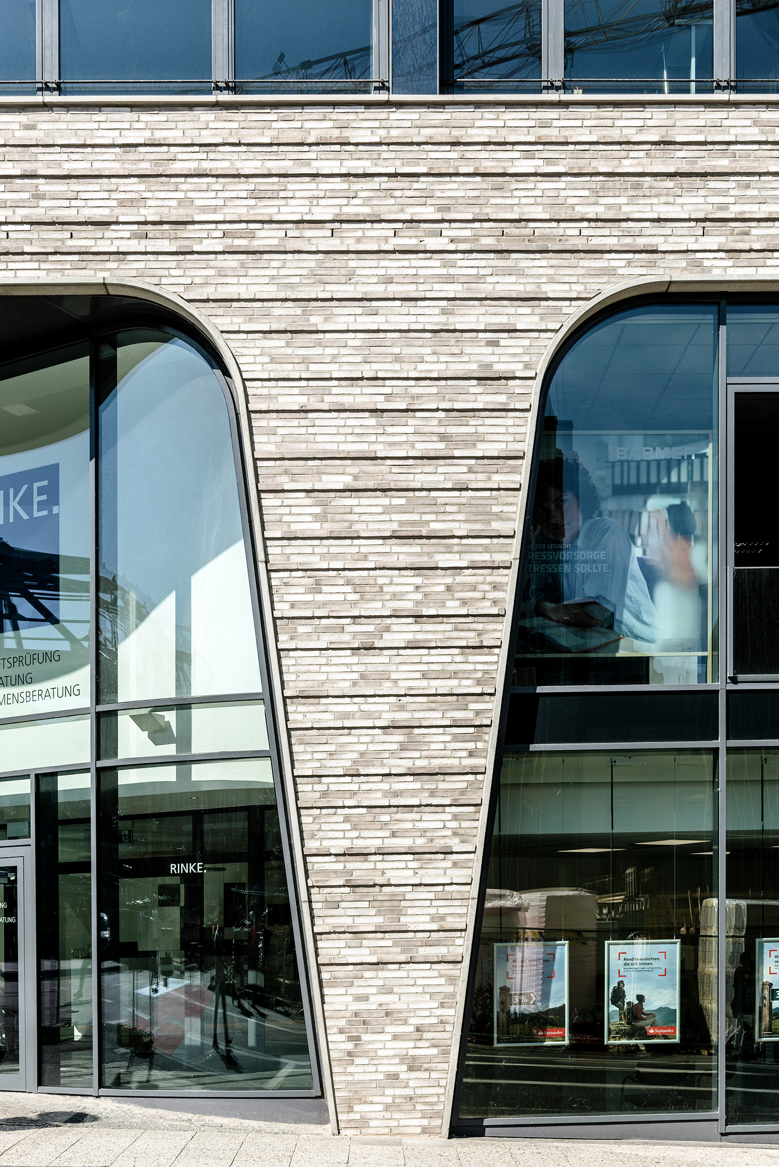 Moderne Gebäudefassade mit hellen Backsteinmauern und großen, einzigartig geformten dreieckigen Fenstern, die die Außenwelt reflektieren. Das untere Fenster zeigt eine Glastür; das obere Fenster gibt den Blick auf ein Bürogebäude frei, in dem Menschen und Schreibtische zu sehen sind.