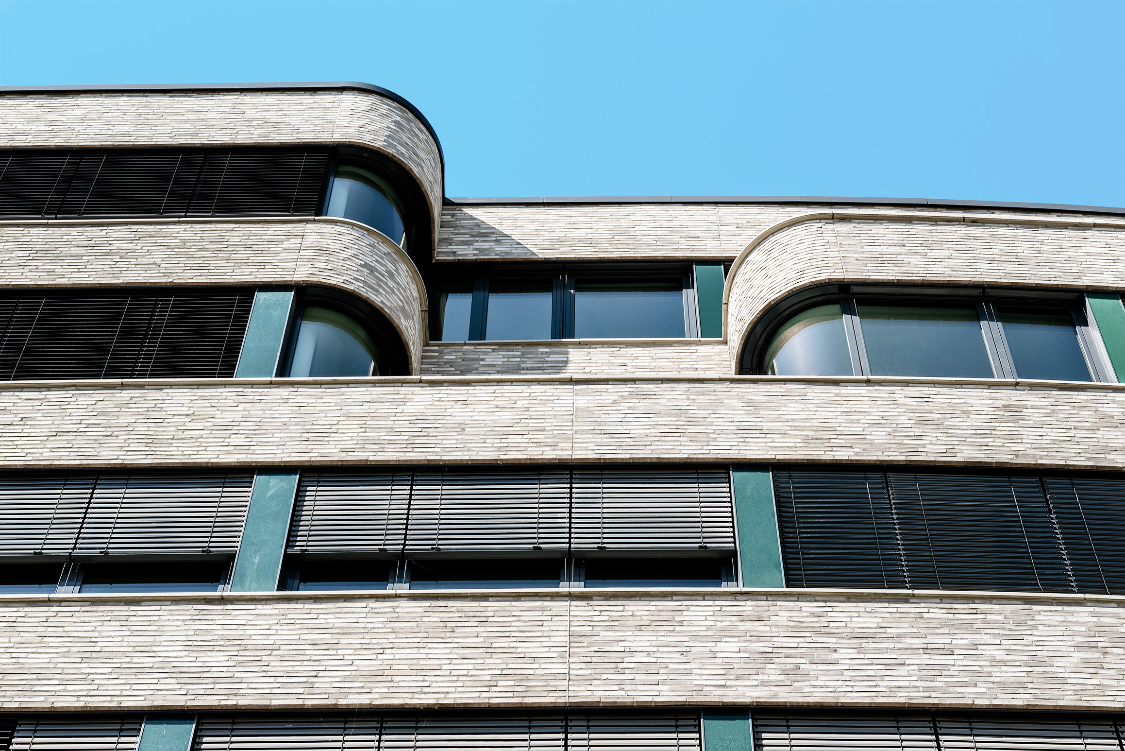 Moderne Gebäudefassade mit geschwungenem Mauerwerk, großen Fenstern und horizontalen schwarzen Jalousien vor einem klaren blauen Himmel.