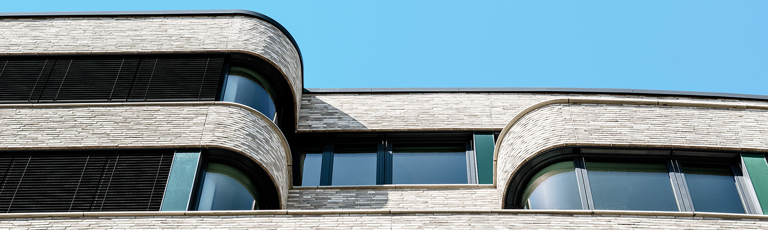 Modernes Gebäude mit geschwungenen, hellen Ziegelwänden und großen Fenstern vor einem strahlend blauen Himmel. Das Design zeichnet sich durch dunkle Fensterrahmen und schlanke, horizontale Linien aus.