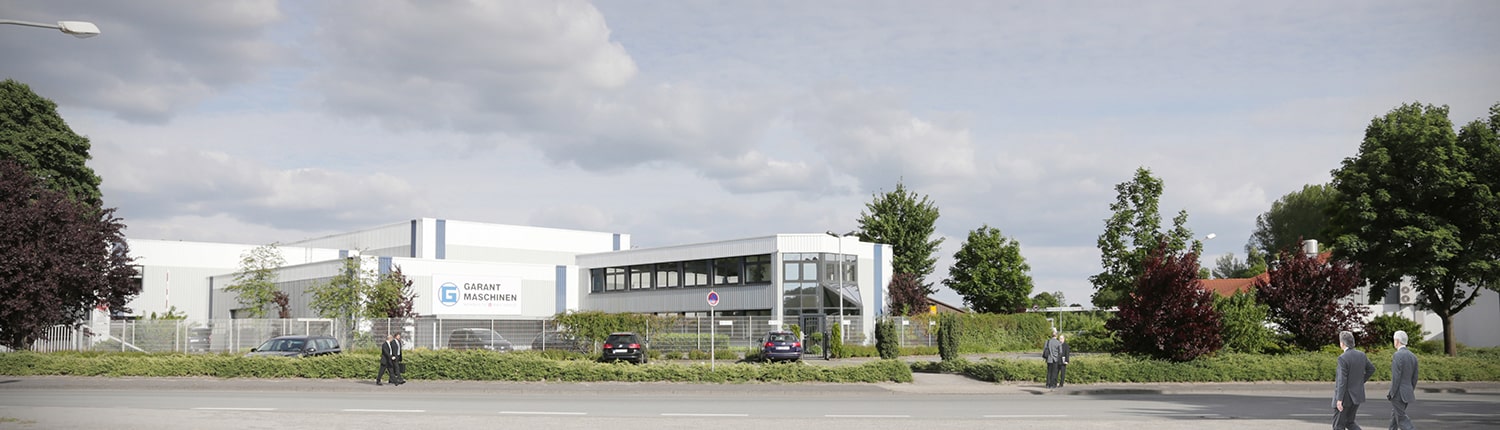 Ein modernes Industriegebäude mit dem Schild „GABBY MIGSCHER“, umgeben von Bäumen und Grünflächen. Mehrere Menschen gehen auf dem Gehweg davor spazieren, und ein paar Autos parken unter einem teilweise bewölkten Himmel in der Nähe eines Maschendrahtzauns.
