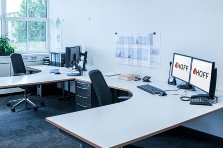 Ein modernes Büro mit zwei L-förmigen Schreibtischen, Bürostühlen, Computern und zwei Monitoren, auf denen HOFF-Inhalte angezeigt werden. An der Wand hängen Diagramme, ein Telefon, Akten und große Fenster sorgen für Tageslicht.