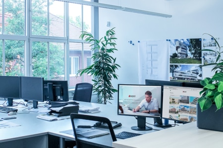 Ein modernes, helles Büro mit mehreren Schreibtischen, Computerbildschirmen, Stühlen und Zimmerpflanzen. Große Fenster lassen Tageslicht herein. An der Wand hängen Architekturzeichnungen und Fotos. Auf einem Computerbildschirm ist ein Mann bei der Arbeit zu sehen.
