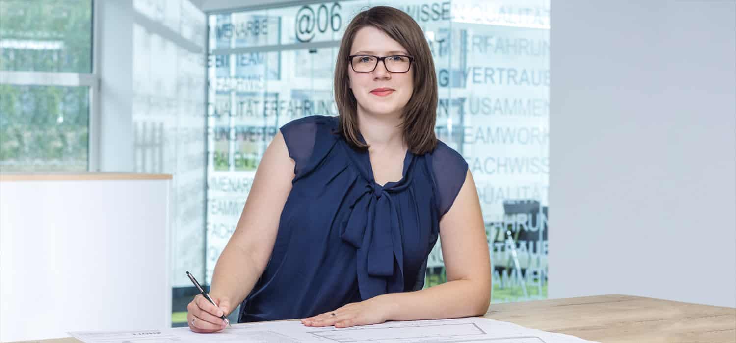 Eine Frau mit Brille und schulterlangem braunem Haar sitzt an einem Schreibtisch, lächelt leicht, hält einen Stift in der Hand und arbeitet an Dokumenten. Sie trägt eine marineblaue Bluse. Im Hintergrund sind Bürofenster zu sehen.
