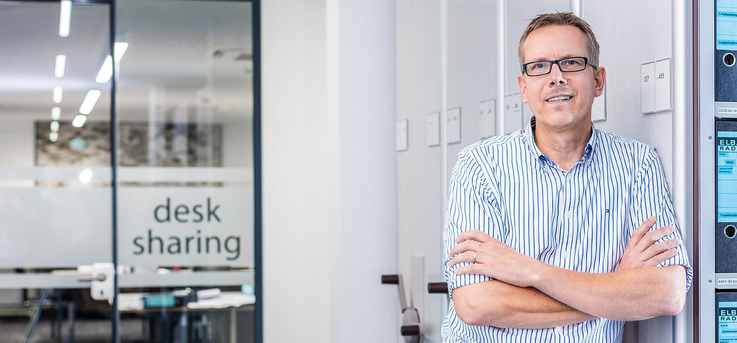Ein Mann mit Brille und gestreiftem Hemd steht lächelnd mit verschränkten Armen neben einer Reihe von Schließfächern in einem modernen Büro. Im Hintergrund ist auf einer Glaswand der Schriftzug „Desk Sharing“ zu sehen.