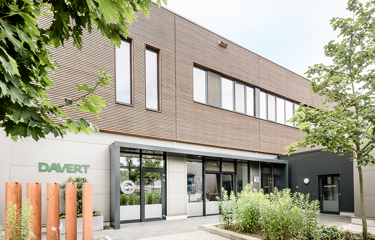 Modernes Gebäude mit braun-weißer Fassade, großen Fenstern und Grünflächen im Vordergrund. Links vom Eingang prangt der Schriftzug DAVERT. Bäume und Pflanzen umgeben den Eingangsbereich.