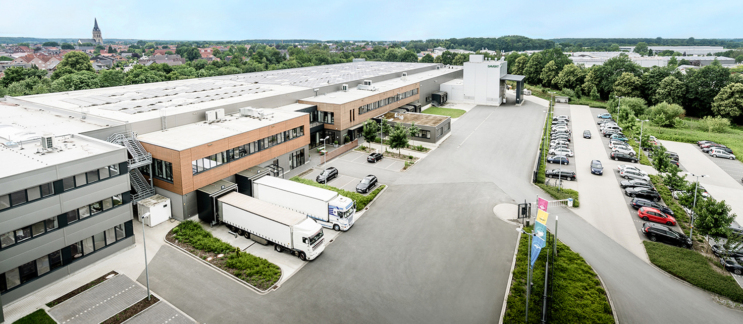 Luftaufnahme eines großen Industriegebäudes mit Laderampen, geparkten LKWs, nahegelegenen Parkplätzen mit Autos, Grünflächen und einer Stadt mit Häusern und einem Kirchturm im Hintergrund.