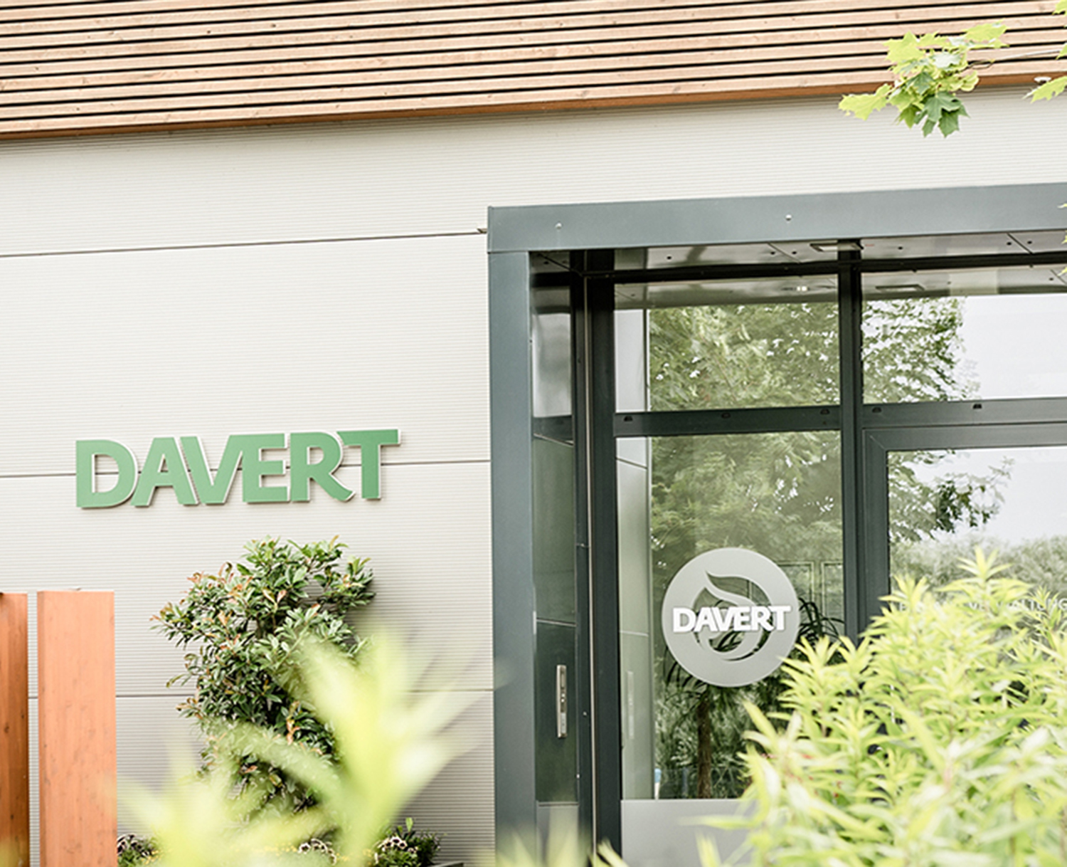 Moderne Gebäudefassade mit grünem DAVERT-Schild an der Wand und einer Glastür mit demselben Logo. Im Vordergrund sind grüne Pflanzen und Bäume zu sehen, die eine natürliche und umweltfreundliche Atmosphäre vermitteln.