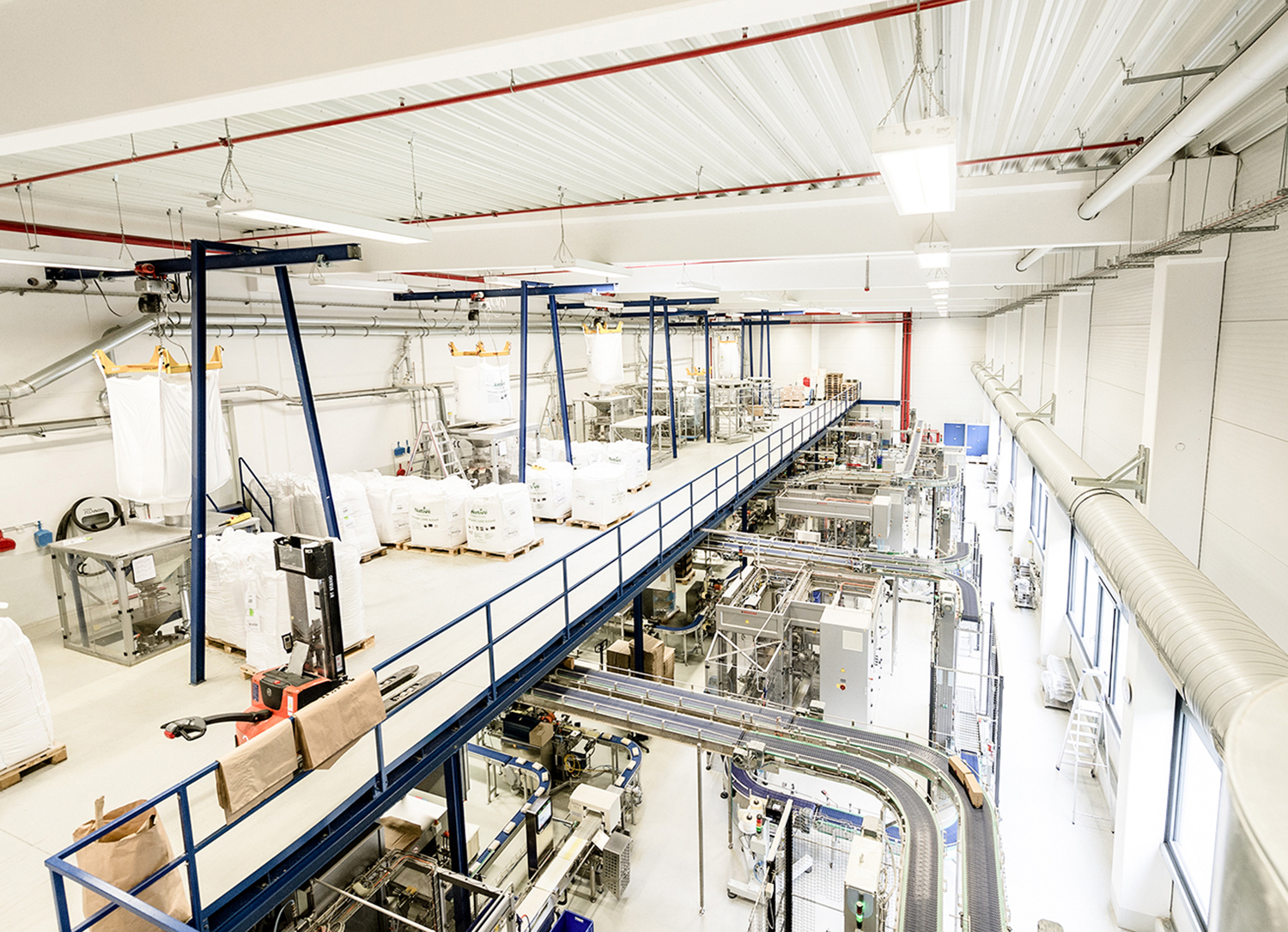 Eine helle, moderne Fabrikhalle mit großen Maschinen, Förderbändern und Säcken voller Rohstoffe. Der Raum ist sauber und ordentlich, mit Metallstegen und Deckenbeleuchtung.