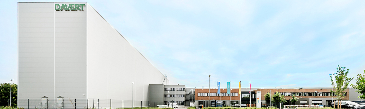 Ein modernes Industriegebäude mit der Aufschrift DAVERT auf einer weißen Fassade, neben einem Komplex aus Bürogebäuden und bunten Flaggen, vor einem blauen Himmel mit vereinzelten Wolken.