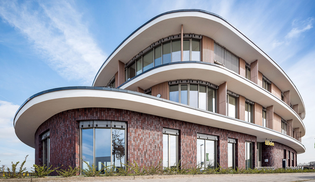 Ein modernes, dreistöckiges Bürogebäude mit geschwungener, geschichteter Architektur, großen Fenstern und einer Ziegelfassade im Untergeschoss. Der Himmel ist klar mit einigen Wolken. Auf dem Gebäude prangt der Schriftzug „Leon“.