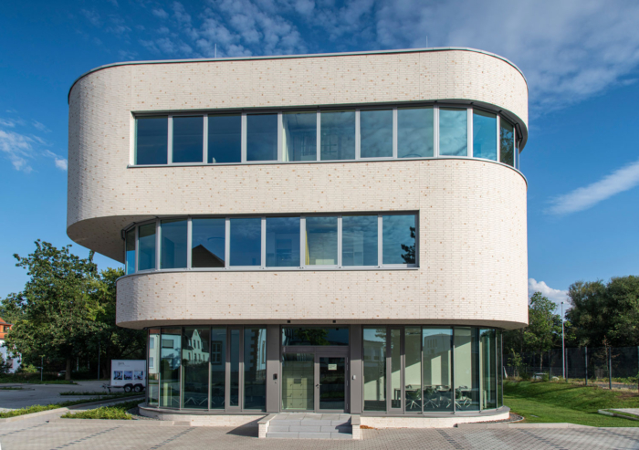 Ein modernes dreistöckiges Bürogebäude mit abgerundeten Kanten, großen Glasfenstern und einer hellen Backsteinfassade vor einem teilweise bewölkten blauen Himmel mit Bäumen und Grün im Hintergrund.