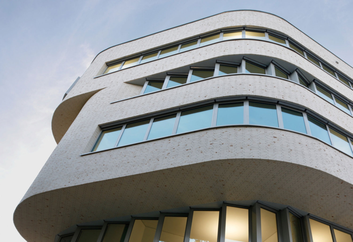 Ein modernes Bürogebäude mit geschwungenen weißen Backsteinwänden und großen horizontalen Fenstern, aus einem niedrigen Winkel vor blauem Himmel fotografiert. Innen sind einige Lichter eingeschaltet und verbreiten ein warmes Licht.