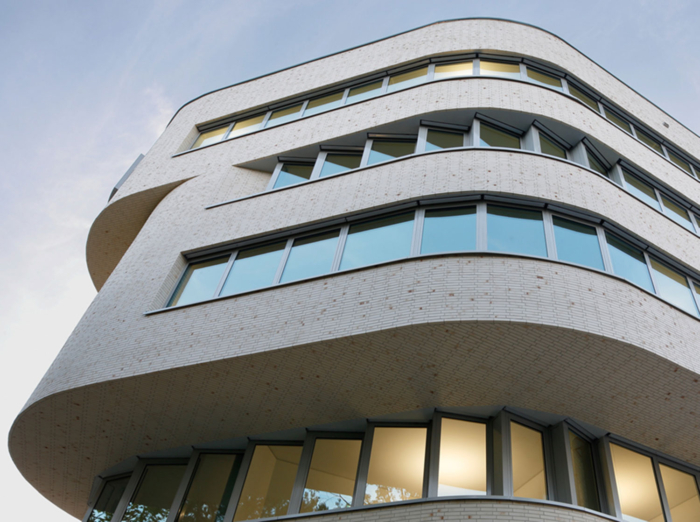 Modernes Bürogebäude mit geschwungener weißer Backsteinfassade, großen Fenstern und mehreren Stockwerken. Die Fenster spiegeln den Himmel wider, und einige Innenlichter sind eingeschaltet. Die Architektur zeichnet sich durch sanfte, abgerundete Linien und ein zeitgenössisches Design aus.