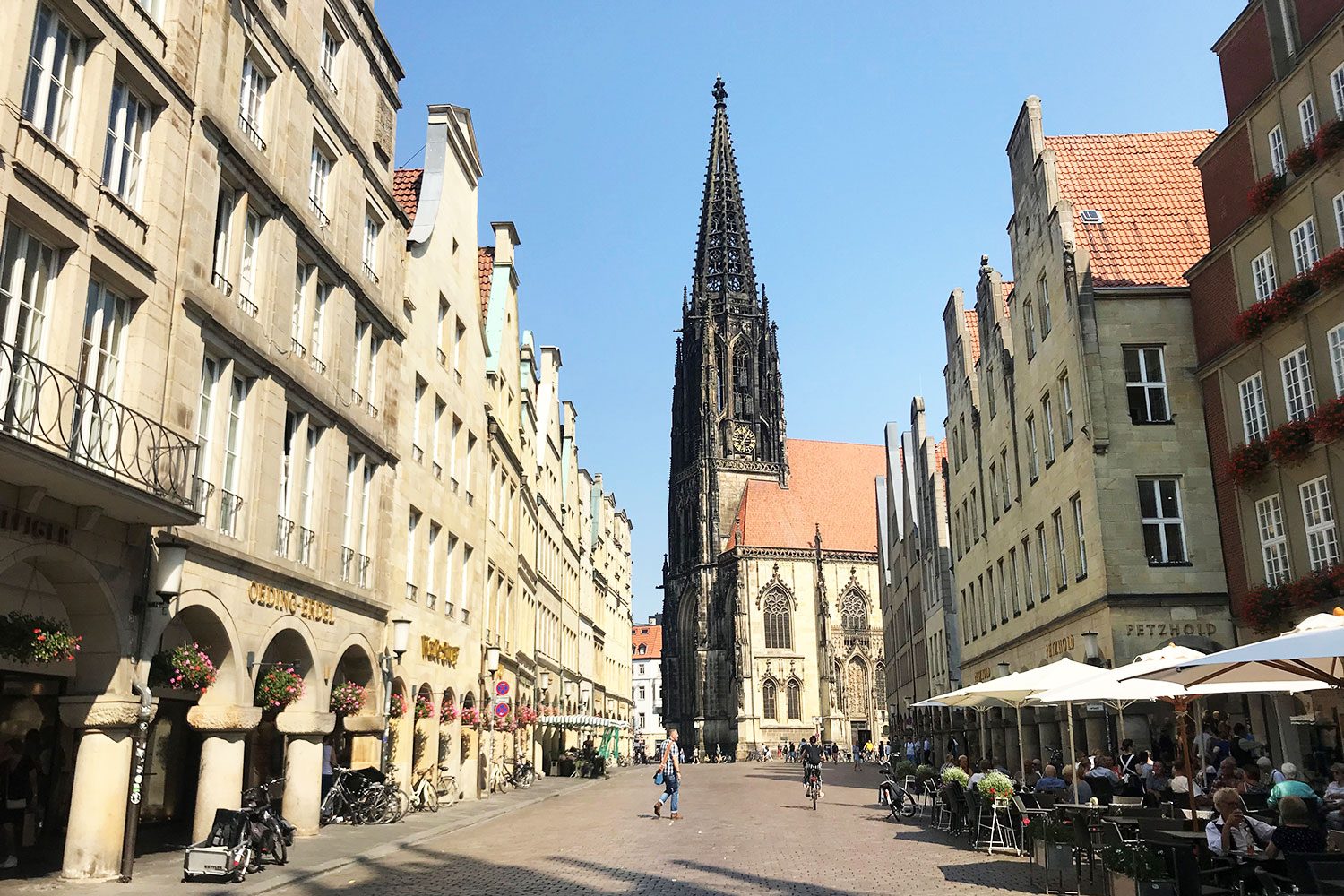 Eine sonnige Straßenszene in einer historischen europäischen Stadt mit hohen, schmalen Gebäuden, Straßencafés und einer großen gotischen Kirche mit einem hohen Turm im Hintergrund. Menschen gehen spazieren und sitzen in den Cafés.