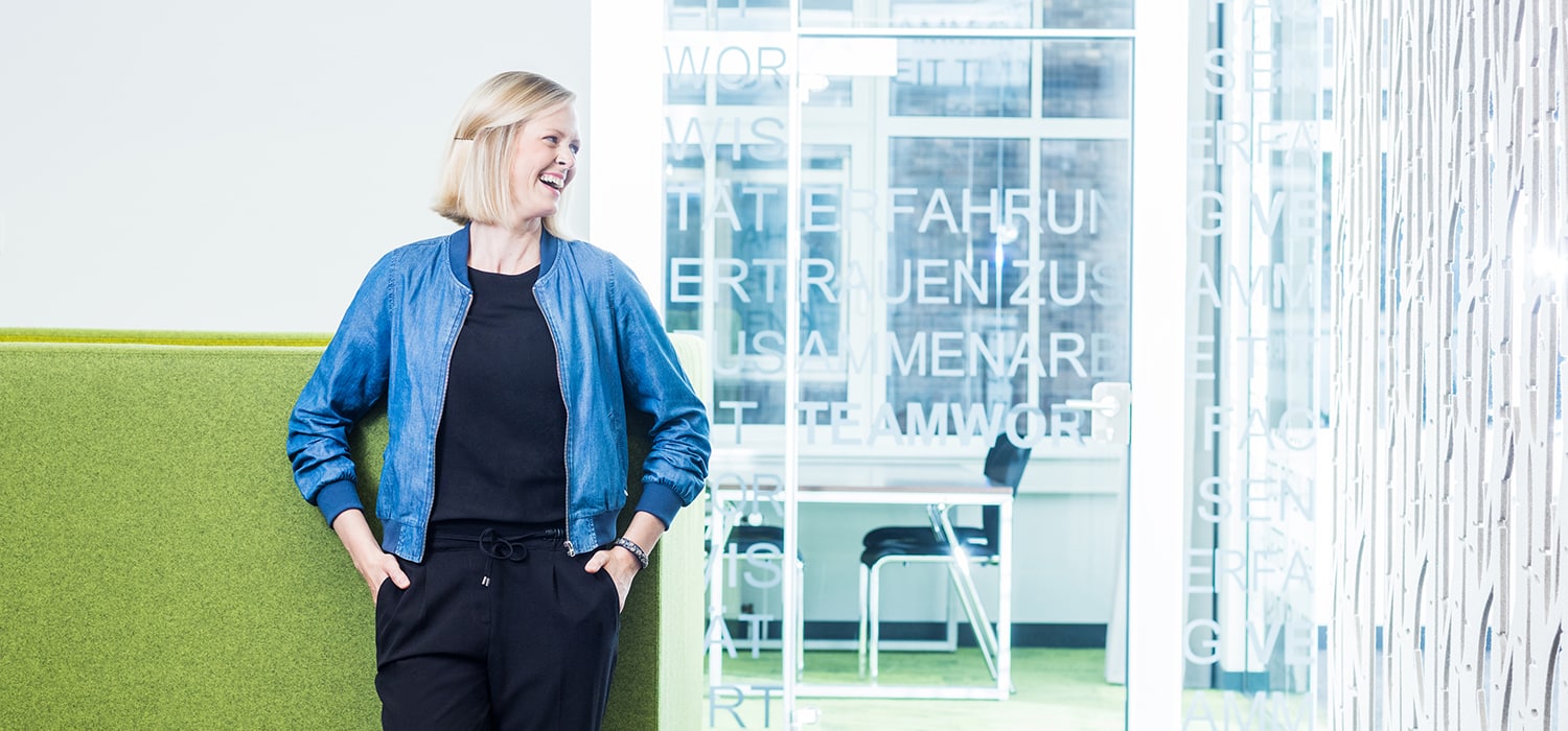 Eine lächelnde Frau mit kurzen blonden Haaren, die eine blaue Jacke und eine schwarze Hose trägt, steht in einem hellen, modernen Büroraum mit grünen Möbeln und Glaswänden mit weißem Text.