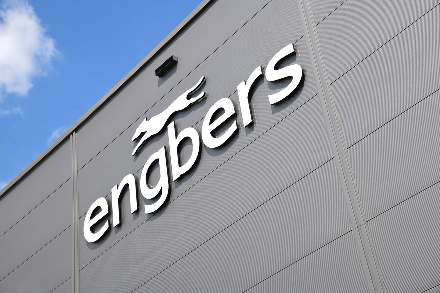 Die Außenfassade eines modernen grauen Gebäudes ziert ein großes weißes Schild mit der Aufschrift „engbers“ in Kleinbuchstaben. Darüber befindet sich ein stilisiertes Logo mit einem laufenden Tier. Zu sehen ist ein blauer Himmel mit einigen Wolken.