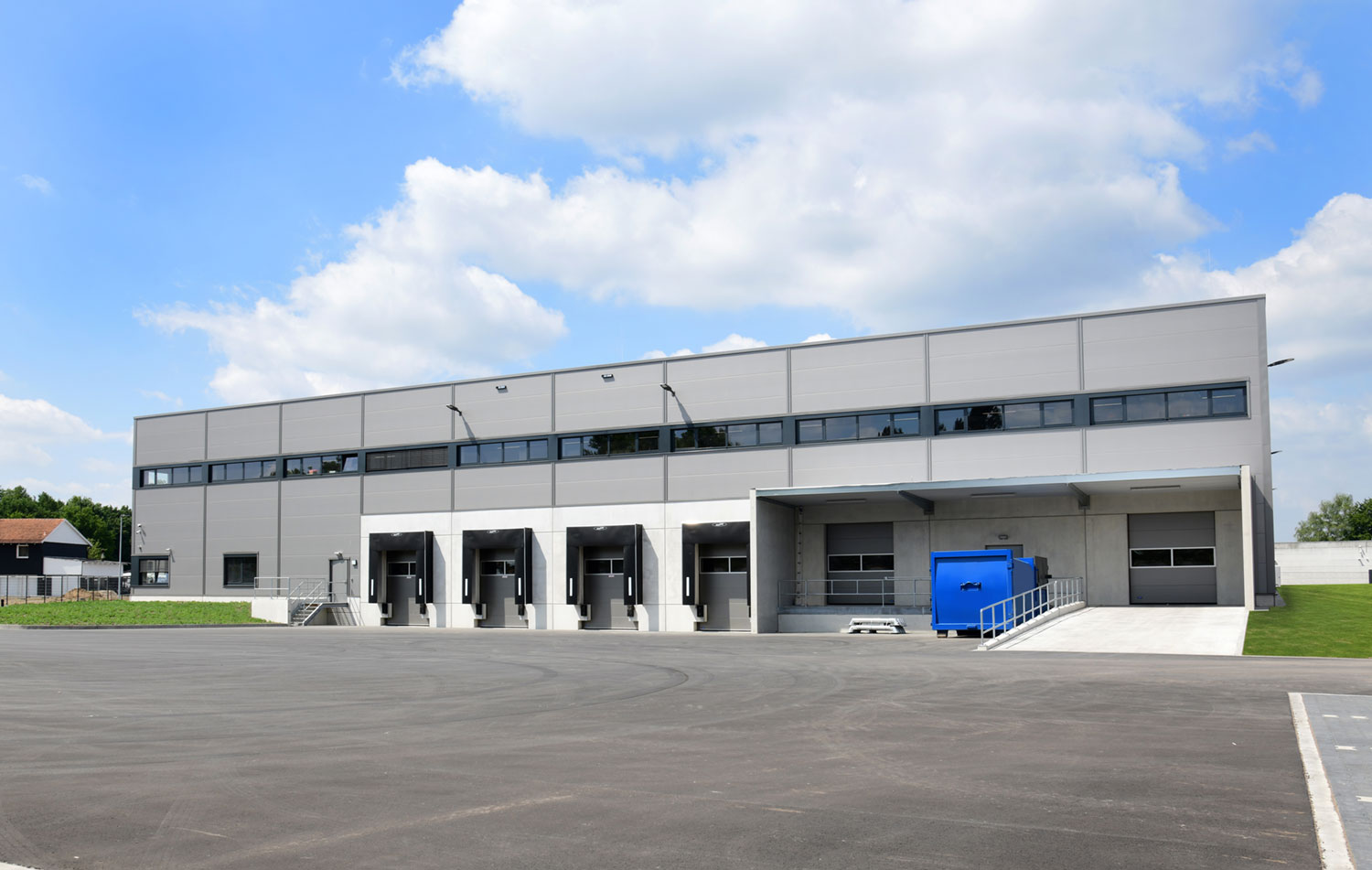 Ein modernes graues Lagerhaus mit mehreren Laderampen, einem blauen Industrietor und einem breiten gepflasterten Bereich davor, vor einem teilweise bewölkten Himmel und grünem Gras.