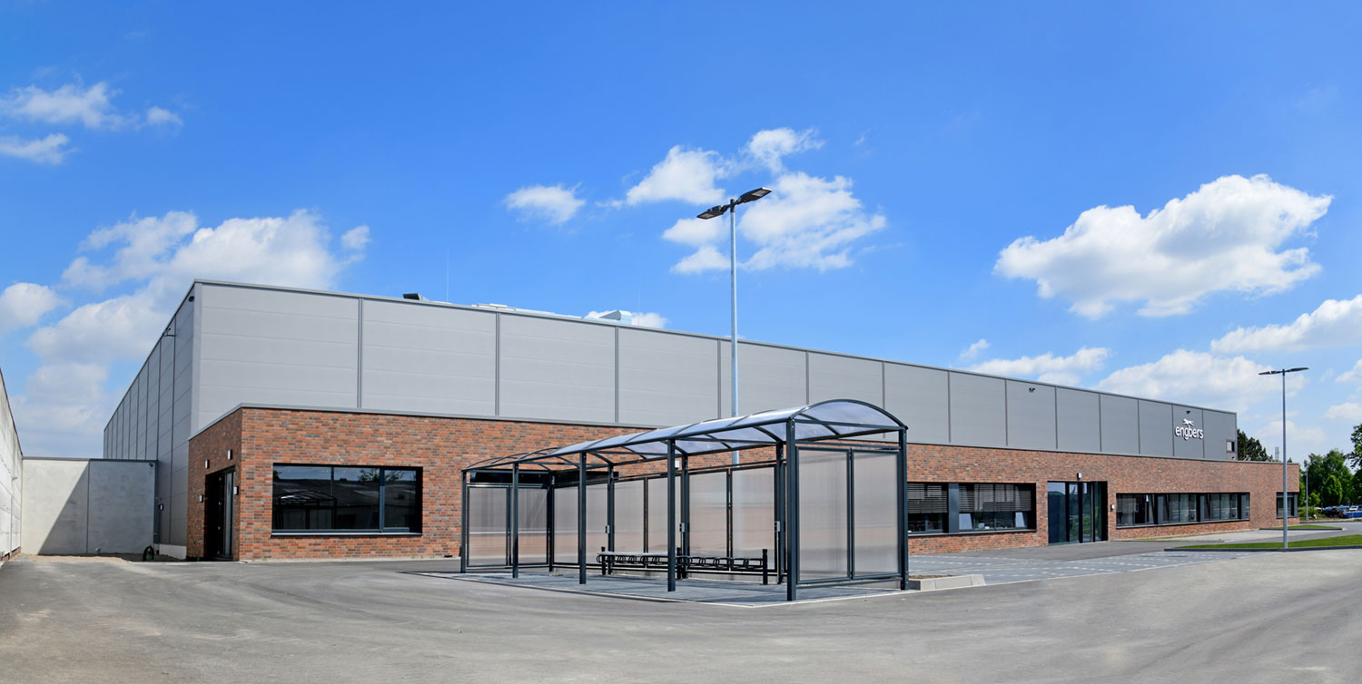 Ein modernes Industriegebäude mit Backstein- und grauer Metallverkleidung, großen Fenstern und einem glasüberdachten Fahrradständer im Freien davor, unter einem blauen Himmel mit vereinzelten Wolken.
