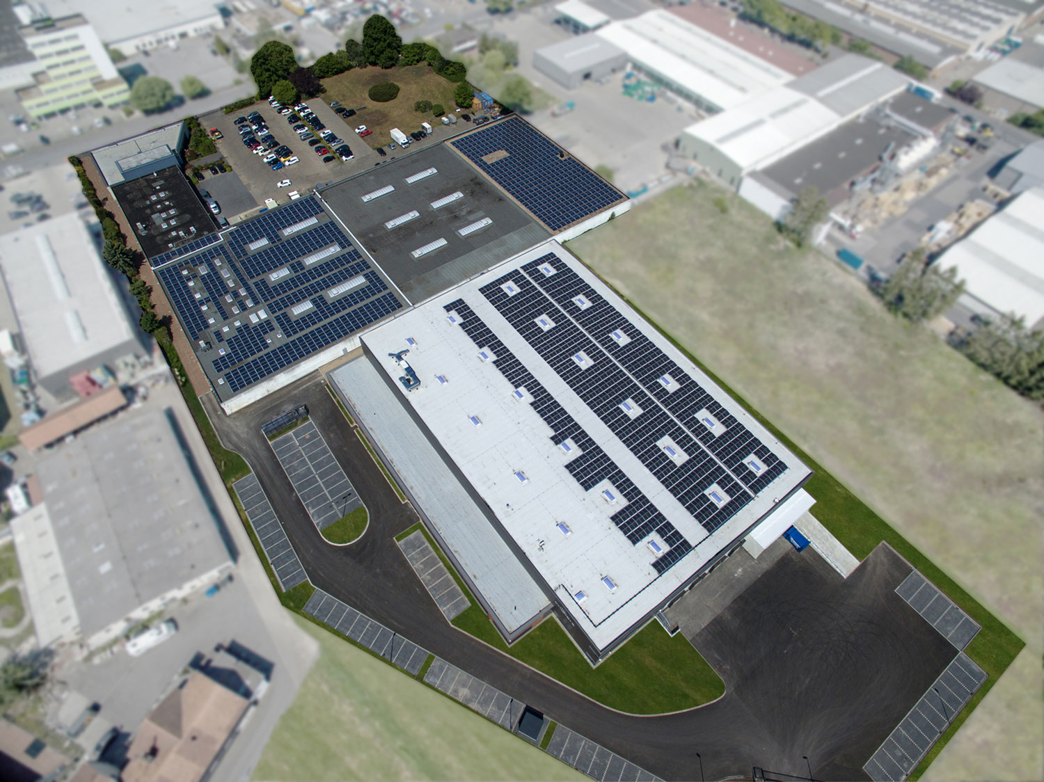 Luftaufnahme einer großen Industrieanlage mit mehreren Gebäuden mit Solarmodulen auf den Dächern, umgeben von Parkplätzen, Straßen und Grünflächen in einem Industriegebiet.