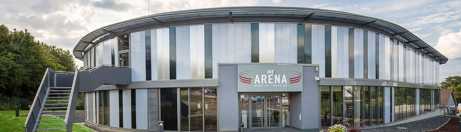 Ein modernes, rundes Sportcenter-Gebäude mit hohen Glasflächen und einem Schild mit der Aufschrift „Die Arena Home of Training“ über dem Eingang. Auf der linken Seite befindet sich eine Außentreppe aus Metall. Der Himmel ist teilweise bewölkt.