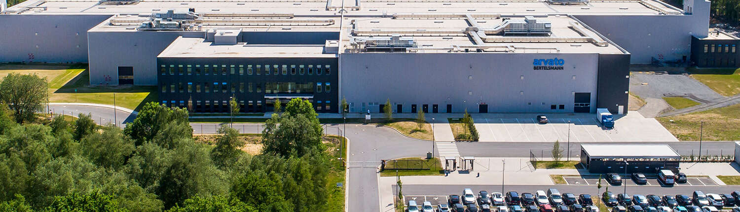 Ein großes industrielles Logistikzentrum mit Arvato Bertelsmann-Beschilderung, umgeben von Grün, einem Parkplatz voller Autos und breiten Straßen, an einem klaren Tag von oben fotografiert.