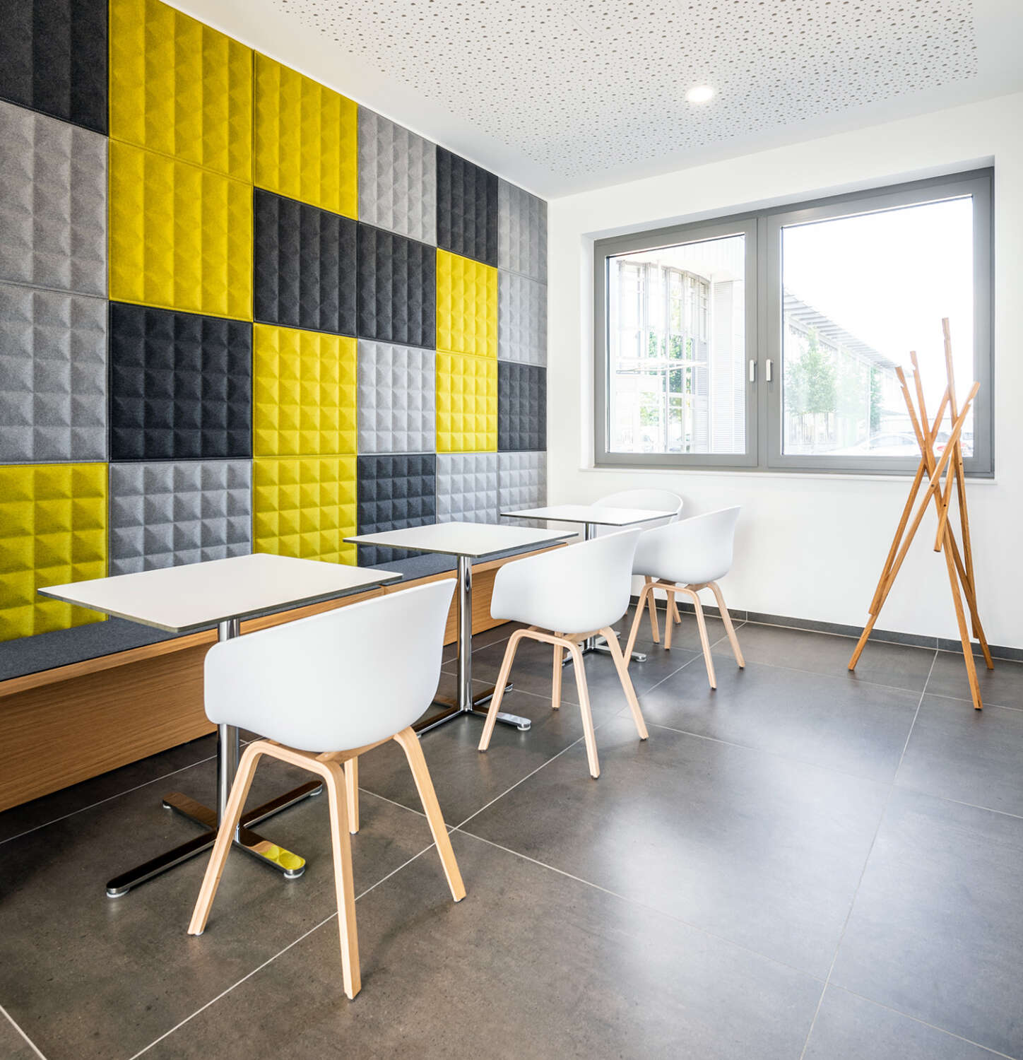 Modernes Bürogebäude mit weißen Stühlen, hellen Holzbeinen, weißen Tischen und einer Wand aus grauen und gelben Akustikplatten. Große Fenster und eine hölzerne Garderobe sorgen für natürliches Licht und erhellen den Raum.