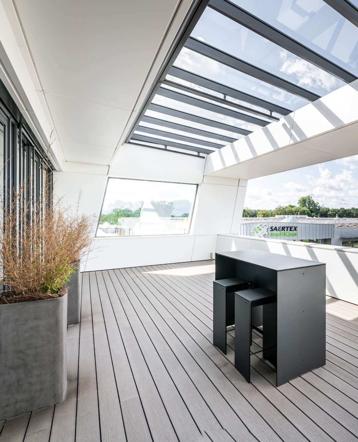 Eine moderne Dachterrasse auf einem Bürogebäude verfügt über helle Holzdielen, einen eleganten schwarzen Tisch mit zwei Hockern, Topfpflanzen und große Glaswände mit einer Glasdecke, die den Raum mit natürlichem Licht durchflutet und Ausblicke auf Bäume und Industriegebäude bietet.