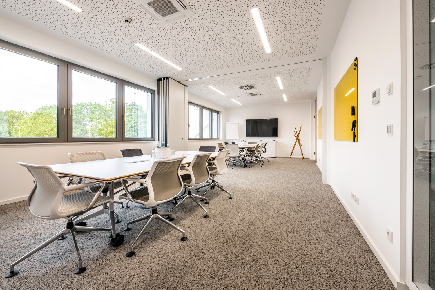 Ein moderner, heller Konferenzraum in einem Bürogebäude mit großen Fenstern, mehreren Tischen und Stühlen, einem an der Wand montierten Fernseher, einer gelben Pinnwand und einer Garderobe. Der leere Raum ist in neutralen Farben und klaren Linien gehalten.