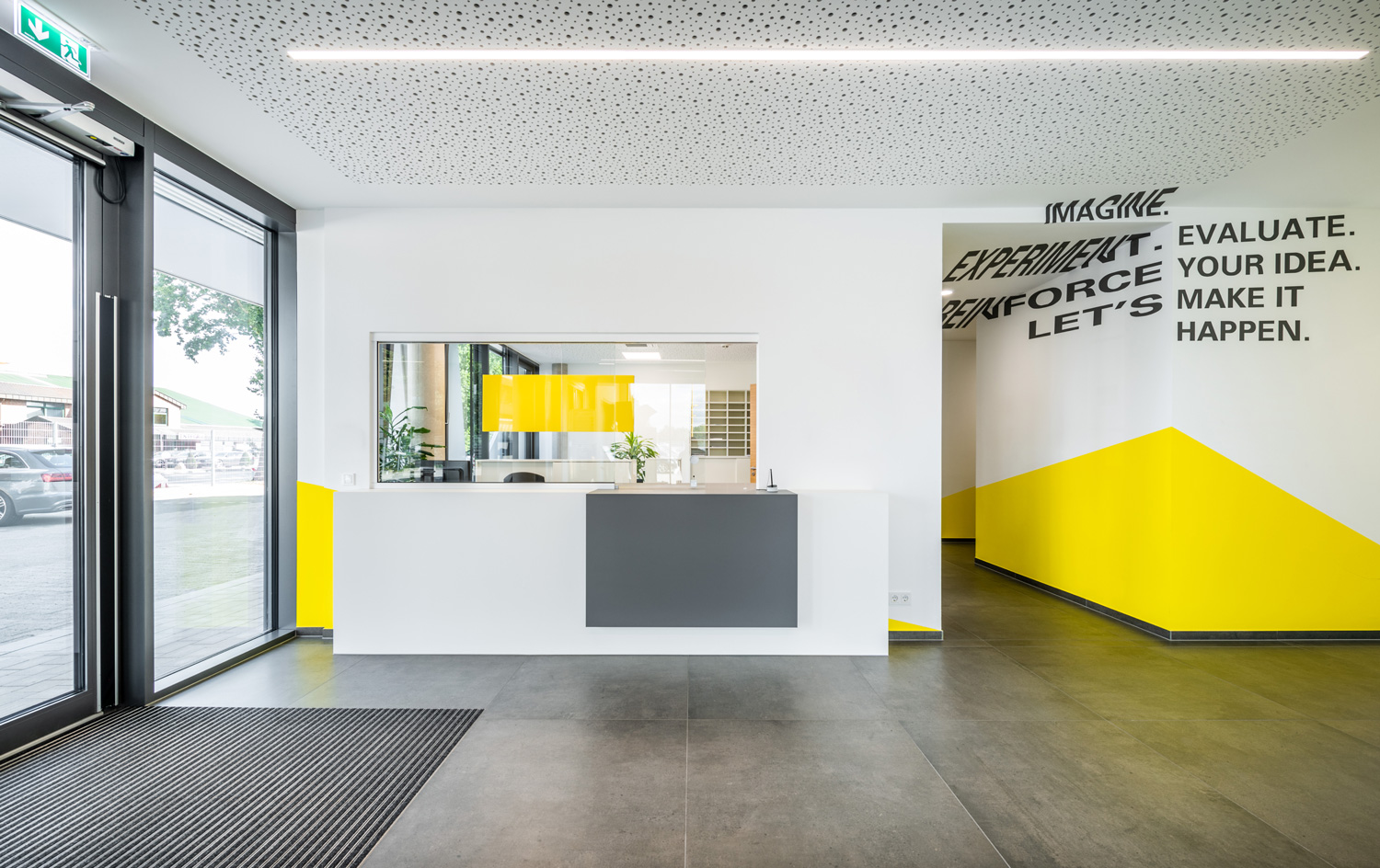 Die Lobby eines modernen Bürogebäudes mit einem weiß-gelben Empfangstresen, großen Fenstern und motivierenden Worten an der Wand: IMAGINE. EXPERIMENT. EVALUATE. REINFORCE. LET’S MAKE IT HAPPEN.