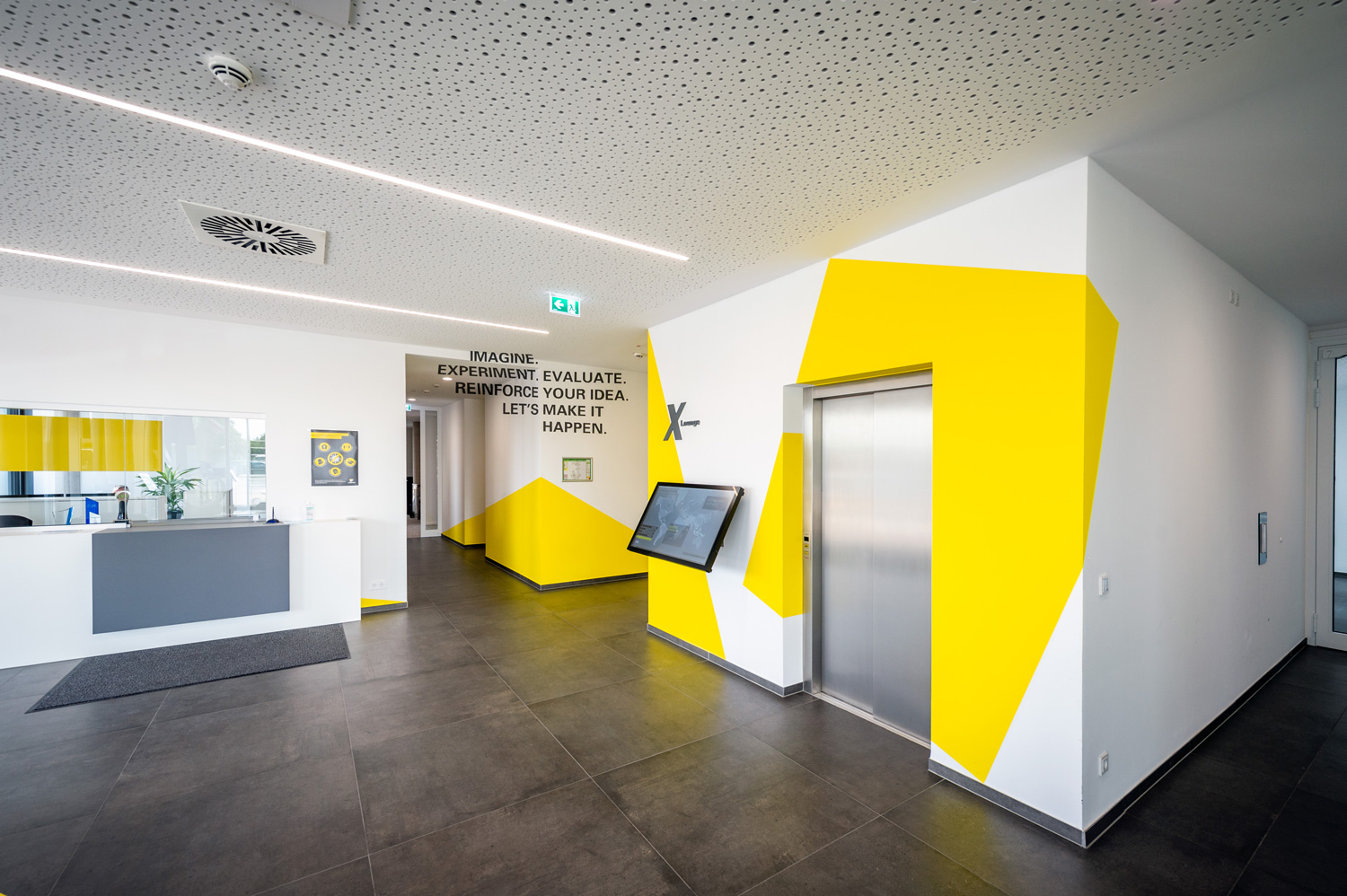 Moderne Lobby eines Bürogebäudes mit weißem Empfangstresen, gelben geometrischen Wandakzenten, Aufzug und Wandbildschirm. Über dem Aufzug hängt ein motivierendes Zitat in diesem hellen und sauberen Raum.
