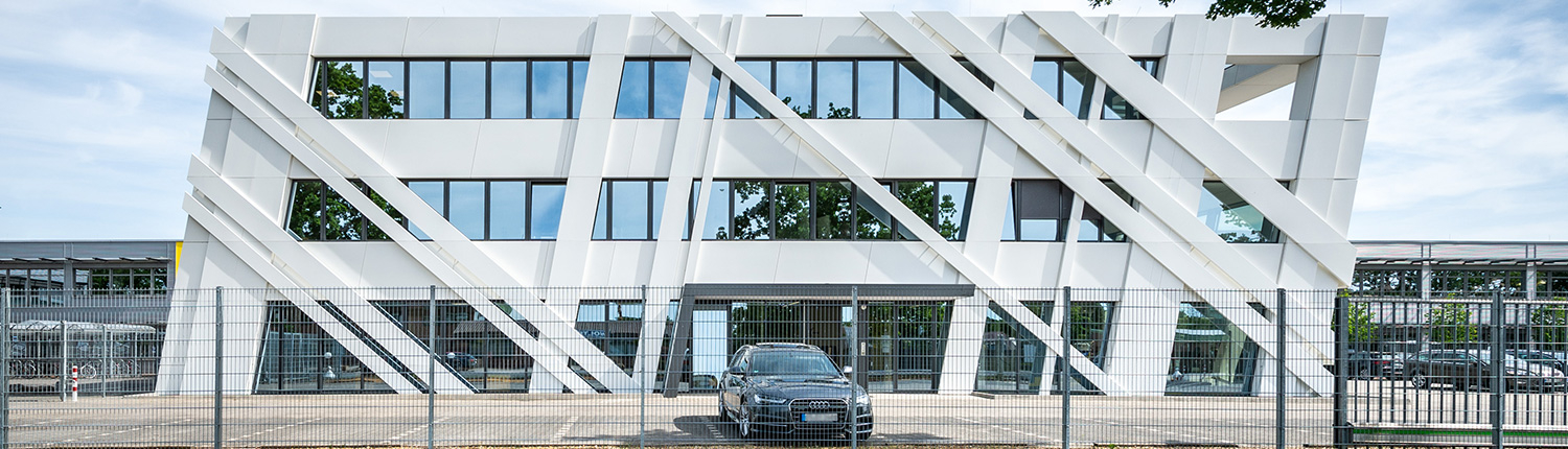 Modernes weißes Bürogebäude mit großen Fenstern und diagonalen Architekturpaneelen an der Fassade, von vorne betrachtet. Ein schwarzes Auto parkt vor dem Gebäude hinter einem Metallzaun. In den Fenstern spiegeln sich Bäume.
