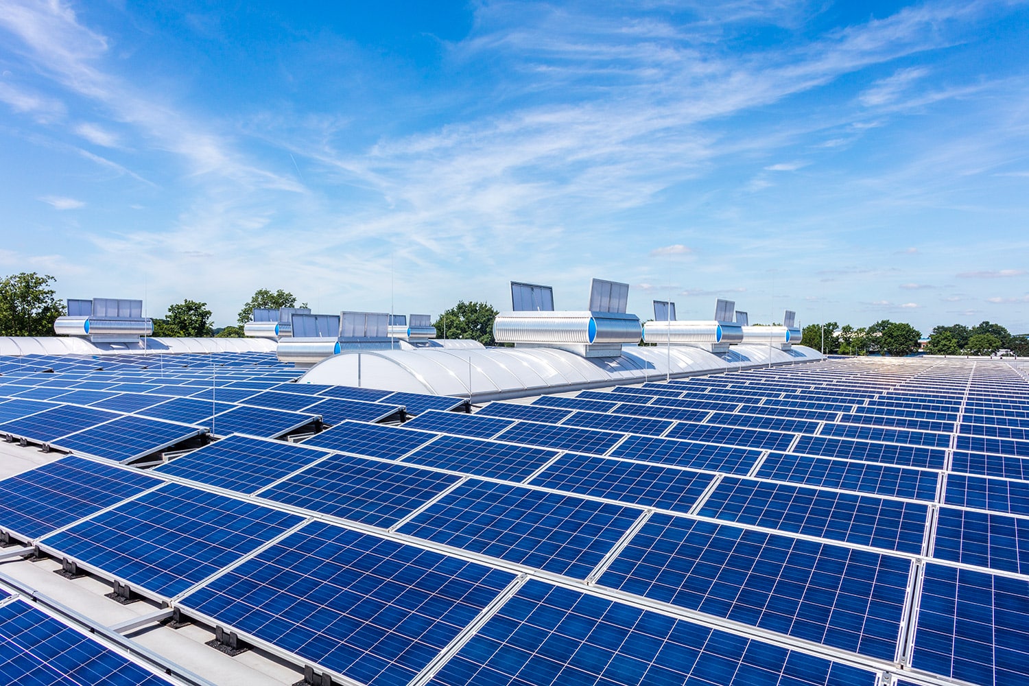 Reihen blauer Solarmodule sind auf einem großen Flachdach unter einem hellen, größtenteils klaren Himmel installiert, im Hintergrund sind ein paar weiße Wolken und einige Bäume zu sehen.
