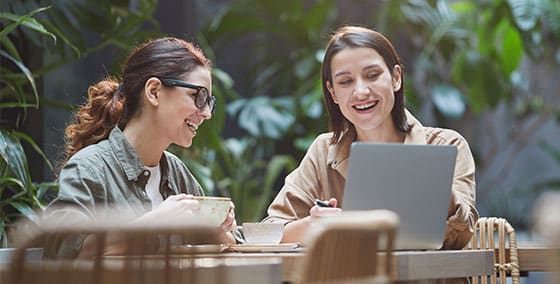 Zwei Frauen sitzen in einem hellen, grünen Innenraum an einem Tisch, lächeln, schauen gemeinsam auf einen Laptop-Bildschirm und genießen ihr Gespräch – ein inspirierender Moment der Freundschaft und des beruflichen Aufstiegs bei einer Tasse Kaffee.