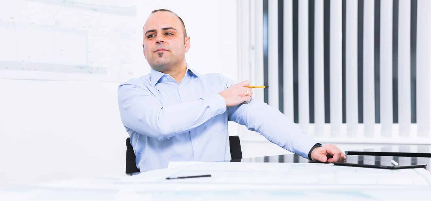 Josef Altun, in einem hellblauen Hemd, sitzt mit Papieren und einem Tablet an einem Schreibtisch, hält einen Stift in der Hand, zeigt über die Schulter und blickt selbstbewusst zur Seite in eine helle Büroumgebung.