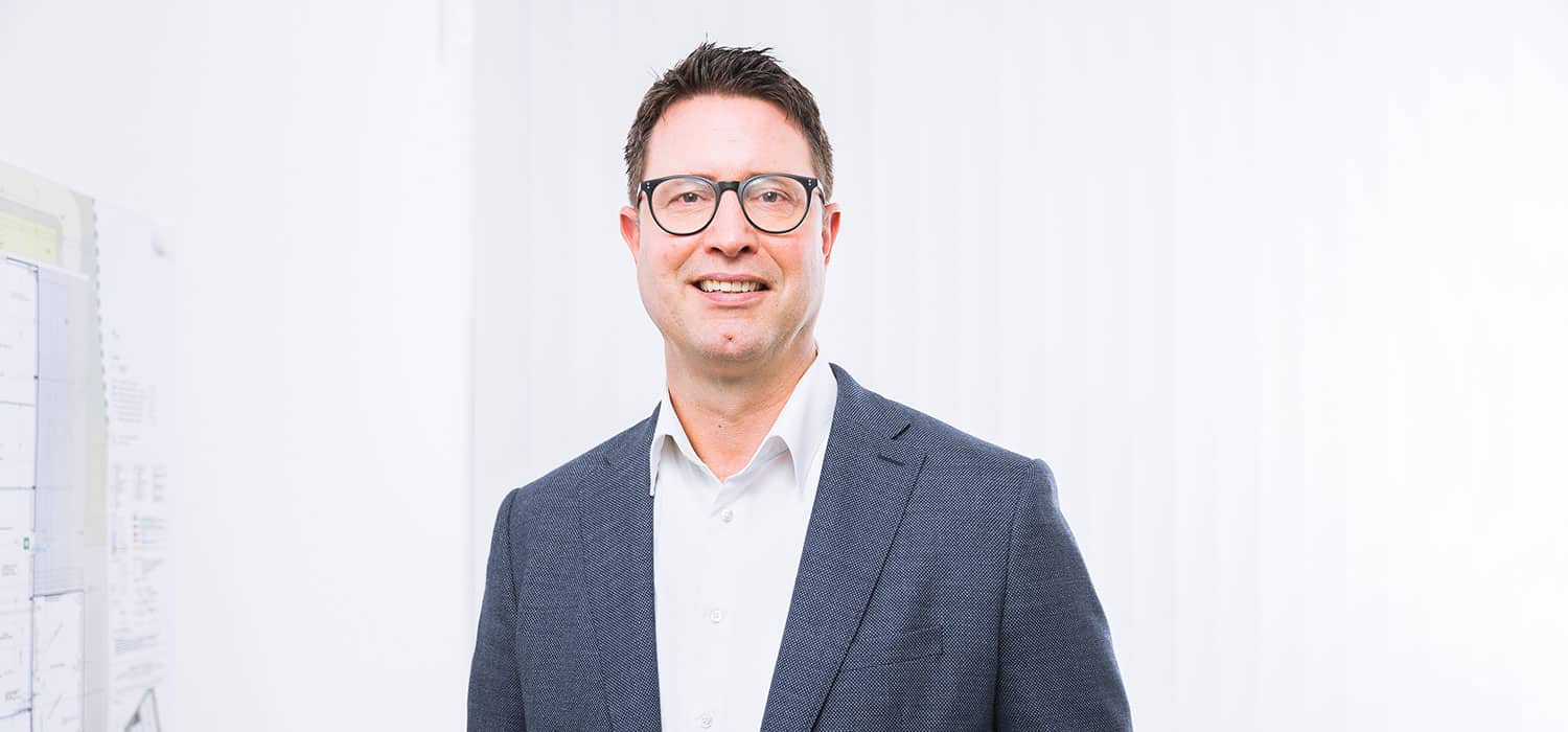 Stefan Bobermin, ein Mann mit kurzen dunklen Haaren und Brille, der ein weißes Hemd und einen dunklen Blazer trägt, steht lächelnd in einem hellen, weißen Büroraum.