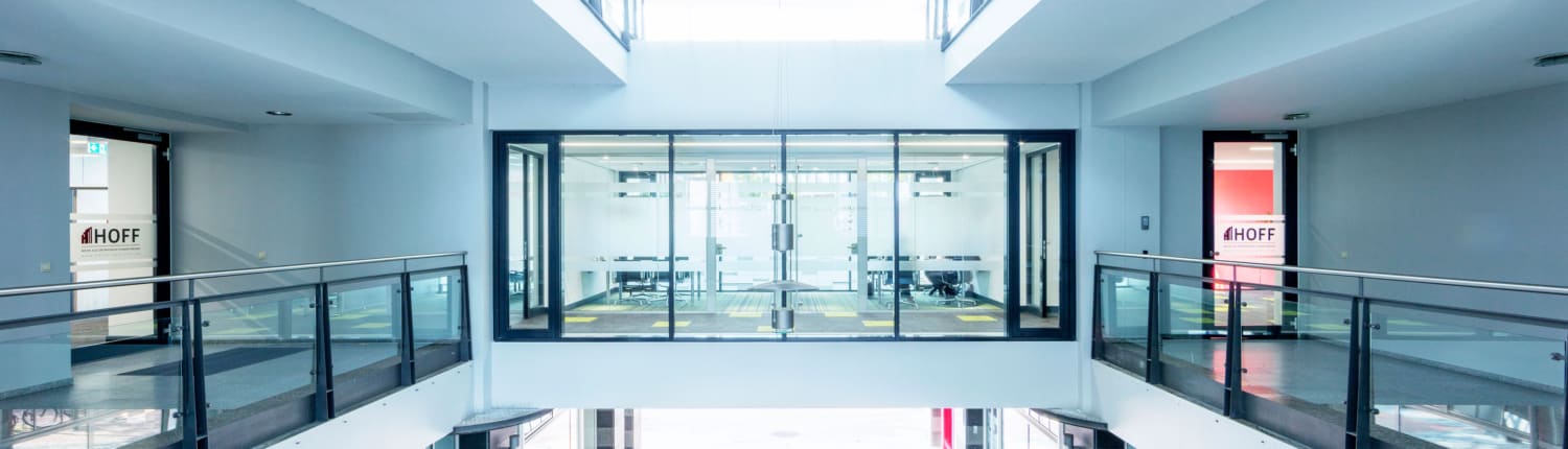 Ein moderner Büroflur mit Glasgeländer und einem verglasten Besprechungsraum in der Mitte. Helles Tageslicht strömt von oben herein und erhellt den klaren, minimalistischen Raum. Auf beiden Seiten stehen Schilder mit der Aufschrift „HOFF“.