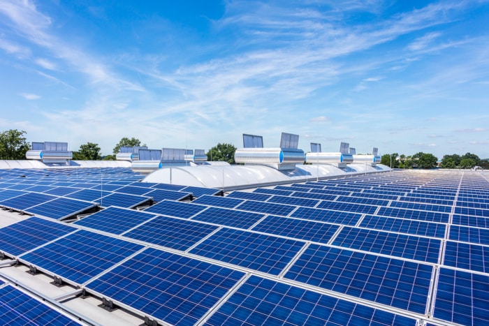 Reihen blauer Solarmodule bedecken ein großes Dach unter einem hellen, teilweise bewölkten Himmel und reflektieren das Sonnenlicht – ein beeindruckendes Beispiel für nachhaltiges Bauen mit sauberer Energie. Im Hintergrund sind einige Bäume zu sehen.