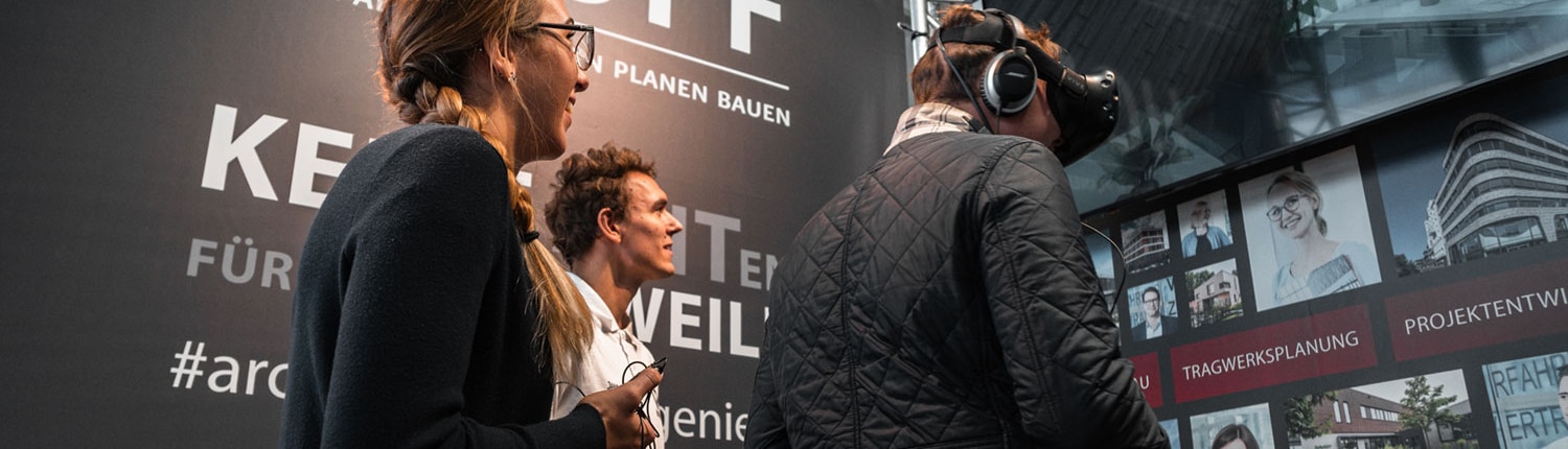 Drei Personen, vermutlich Campus-Studierende, interagieren an einem Messestand mit HOFF-Beschilderung. Eine Person nutzt eine Virtual-Reality-Brille, während die anderen lächelnd zuschauen. Im Hintergrund und an der Wand sind Architekturbilder zu sehen.