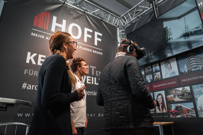 Drei Personen, vermutlich Campus-Studierende, interagieren an einem Messestand mit HOFF-Beschilderung. Eine Person nutzt eine Virtual-Reality-Brille, während die anderen lächelnd zuschauen. Im Hintergrund und an der Wand sind Architekturbilder zu sehen.