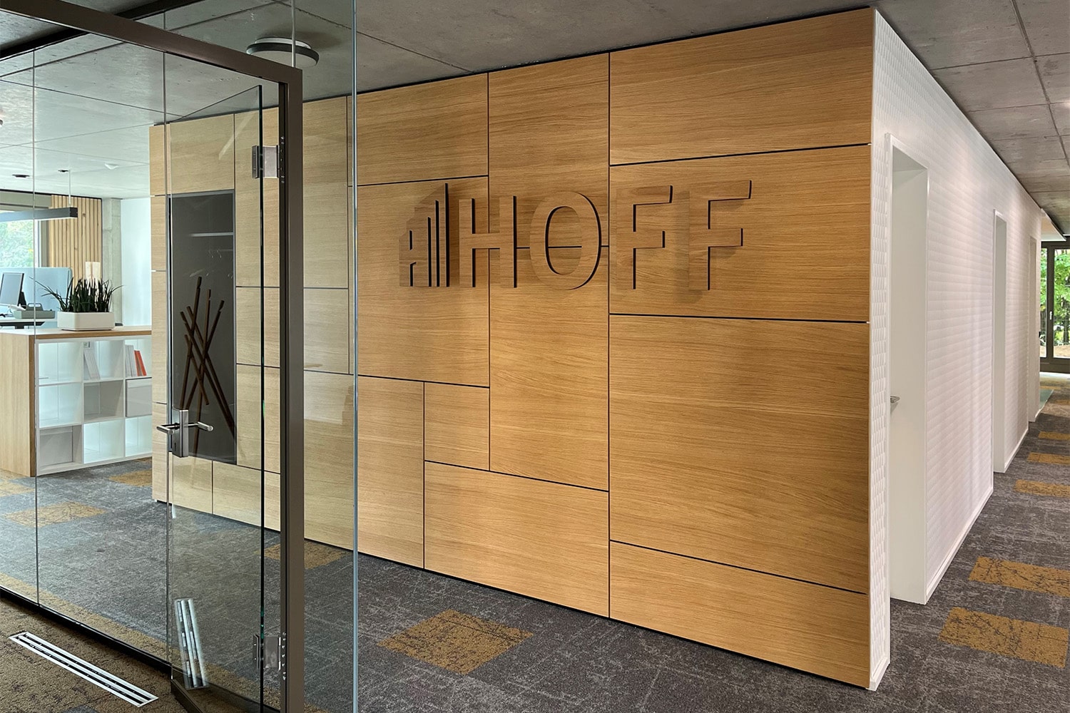 Moderner Büroeingang mit Glastür, holzgetäfelter Wand mit dem Wort HOFF in großen Buchstaben und einem im Hintergrund sichtbaren Empfangsbereich mit Pflanzen und einem weißen Tresen.