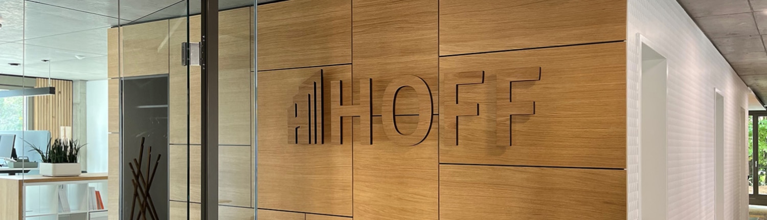 Ein modernes Interieur des Büro Münster mit Holzwandpaneelen, auf denen das Logo und der Name HOFF in großen Buchstaben zu sehen sind, Glastüren und im Hintergrund sind zeitgenössische Möbel zu sehen.